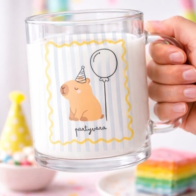 Caneca de vidro com desenho de urso de festa e balão, cheia de leite, numa mesa com decoração de festa.