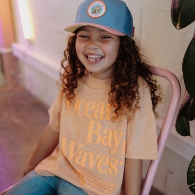 Criança sorridente com t-shirt bege e boné azul claro sentada numa cadeira rosa.