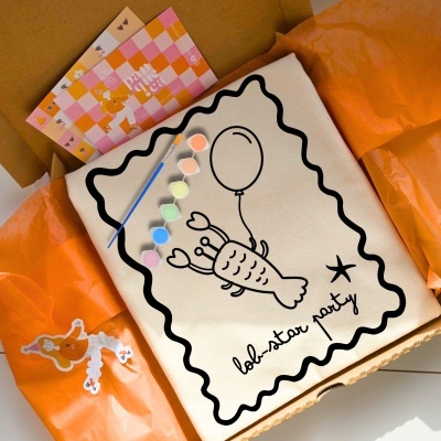 Kit de pintura em tela com desenho de lagosta, pincel e tintas coloridas em caixa com papel laranja
