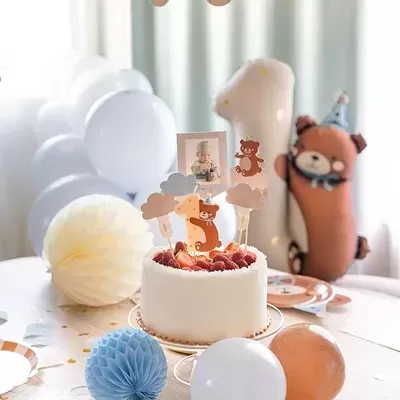 Mesa de festa decorada com bolo branco, balões e enfeites, incluindo um boneco de peluche castanho com chapéu de festa.