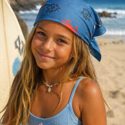 Menina com top de bikini azul claro e lenço azul com desenhos, em praia com prancha de surf ao fundo