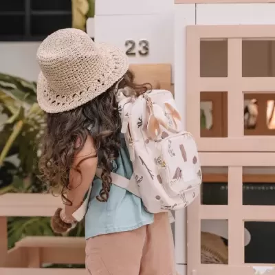 Menina com chapéu de crochet e mochila branca com estampado junto a uma casa de madeira bege e branca.
