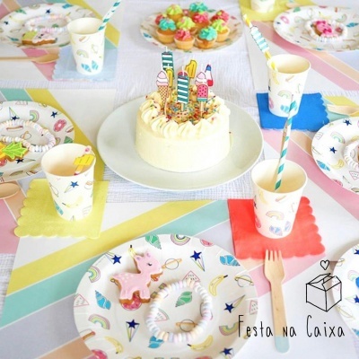 Mesa decorada para festa com pratos e copos coloridos, bolo branco com velas de aniversário, utensílios e padrão de unicórnios e estrelas