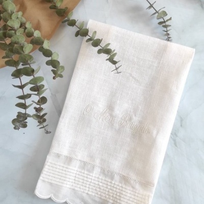 toalha branca bordada com detalhes e ramos verdes sobre fundo branco marmorizado