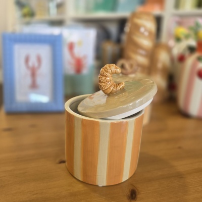 Pote de cerâmica laranja às riscas com tampa bege e pega em forma de croissant sobre mesa de madeira.