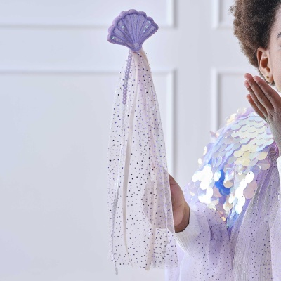 Varinha com cabo de concha roxa e tecido branco com glitter, segurada por criança com camisa lilás de lantejoulas