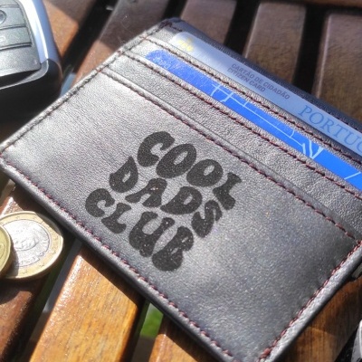 Carteira castanha escura de pele com texto COOL DADS CLUB sobre banco de madeira