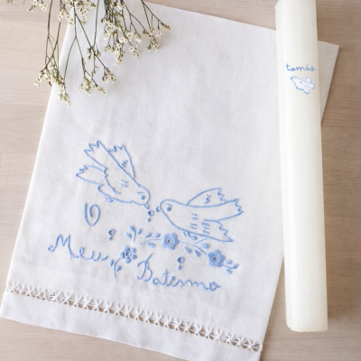 Toalha bordada branca com padrão de pássaros e vela branca decorada, em madeira clara.