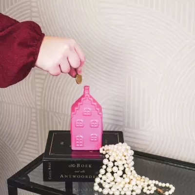 Mealheiro rosa em forma de casa com moeda, sobre livros e colar de pérolas, fundo cinza texturado.