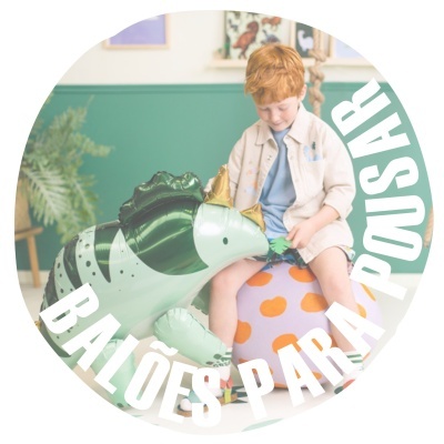 Menino sentado numa bola laranja e branca ao lado de balão verde de dinossauro em ambiente interior