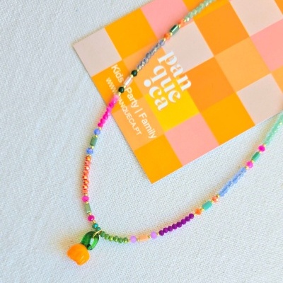 Colar colorido de contas com pendente de flor laranja e conta verde sobre cartão quadriculado com texto.
