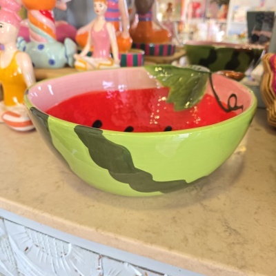 Taça cerâmica pintada com padrão de melancia verde e vermelho, em balcão com figuras coloridas ao fundo