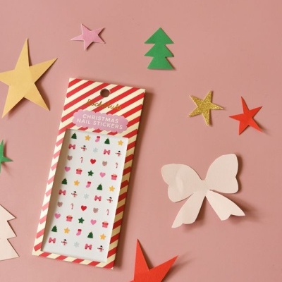 Pacote de autocolantes para unhas de Natal numa superfície cor de rosa com recortes de papel colorido ao redor.
