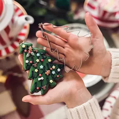 Cookie árvore de Natal decorado com molde metálico, mãos a segurá-los, fundo com chávena e bule às riscas vermelhas e brancas.