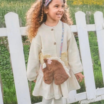 Menina com casaco bege e mala em forma de cavalo peludo castanho, com bandana azul, ao ar livre junto a cerca branca.