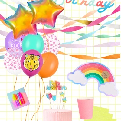 Decoração de festa colorida com balões, bolo rosa, velas, copo e guardanapos.