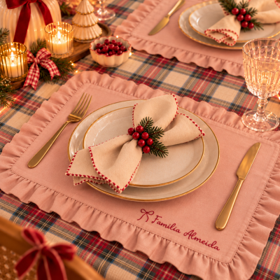 Mesa posta com toalha xadrez, individuais rosas com babados, pratos brancos, talheres dourados, guardanapos decorados e velas acesas.
