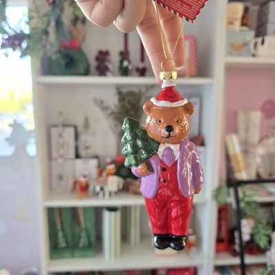 Enfeite de Natal em vidro com urso vestindo casaco lilás e calças vermelhas segurando árvore de Natal verde.