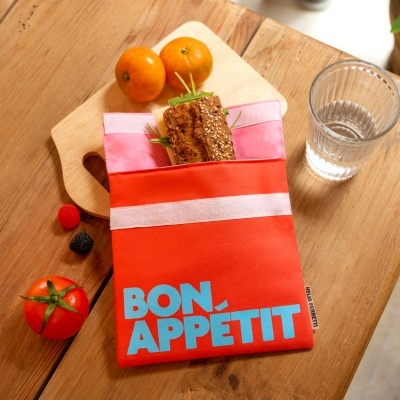 Saco reutilizável vermelho com texto 'BON APPÉTIT' contendo sanduíche, ao lado frutas e copo de água