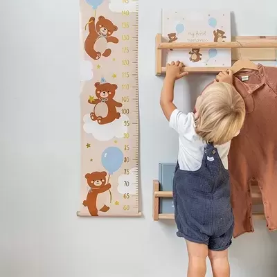 Régua de crescimento para crianças com ursos e balões na parede ao lado de menino vestido com jardineira azul.