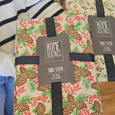 Toalha de mesa com padrão de pinhas, folhas e bagas embaladas com etiqueta preta.