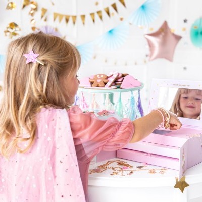 Cajita de madeira rosada com espelho e estrelas douradas numa mesa branca com menina loira e decorações de festa ao fundo