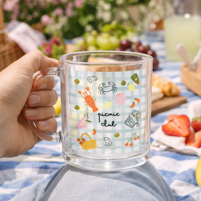 Caneca de vidro com decoração colorida e texto picnic club em ambiente de piquenique