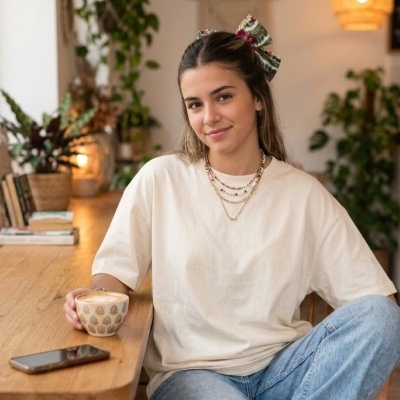 Mulher jovem sentada com t-shirt branca e jeans segurando chávena com padrão geométrico num ambiente interior com plantas