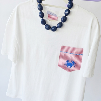 T-shirt branca com bolso vermelho listrado e caranguejo azul pendurada com colar azul e chapéu de palha com lenço vermelho