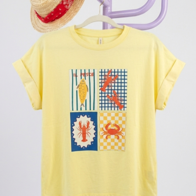 T-shirt amarela com estampas de peixe e crustáceos pendurada num cabideiro com chapéu de palha.