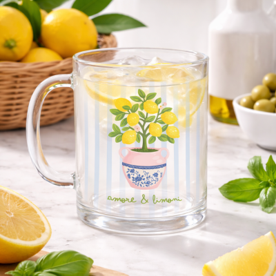 Caneca de vidro transparente com ilustração de limoeiro e texto amore & limoni contendo bebida com fatias de limão e gelo numa mesa com limões, manjericão, azeitonas e garrafa.
