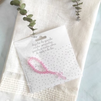 Toalha creme com bainha ondulada e cartão com pulseira rosa num saco plástico sobre a toalha