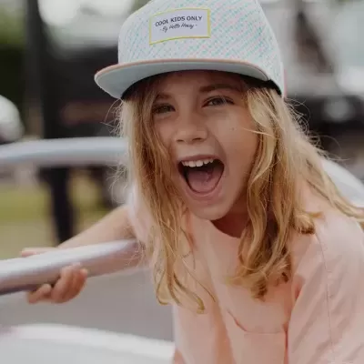 Criança sorridente a usar boné branco com texto e padrão colorido e camiseta rosa claro num parque