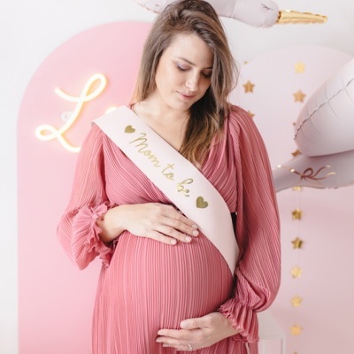 Mulher grávida com vestido rosa e faixa 'Mom to Be' em ambiente decorado com balões e estrelas.