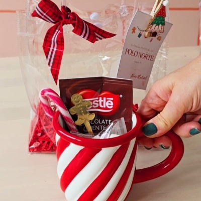 Caneca listrada vermelha e branca com chocolate Nestlé e acessórios em fundo neutro com saco transparente e laço