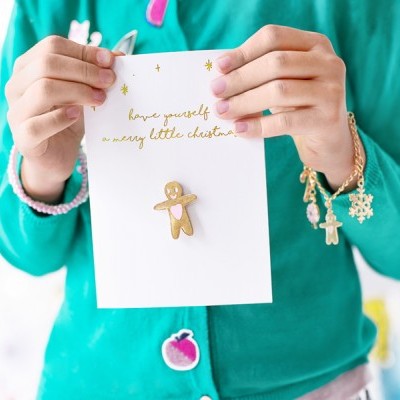 Cartão natalício com mensagem dourada e boneco de gingerbread segurado por pessoa com casaco verde e pulseira