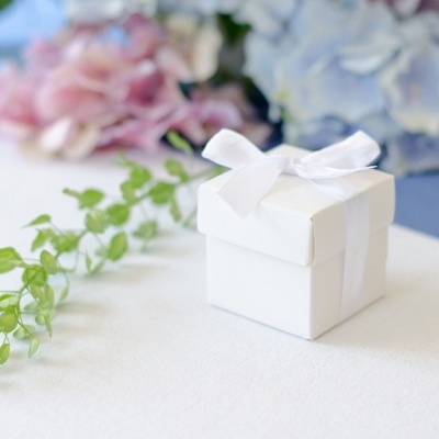 Caixa de presente branca com laço sobre superfície branca e ramos de flores ao fundo