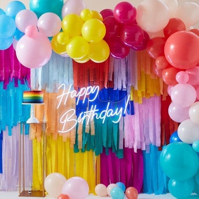 Decoração colorida de festa com balões, franjas de papel e letreiro neon 'Happy Birthday'
