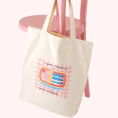 Mala de tecido creme com estampa de lata de sardinhas e texto em letras pretas pendurada numa cadeira rosa