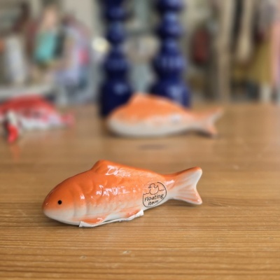 Peixe decorativo de cerâmica laranja e branco sobre mesa de madeira clara