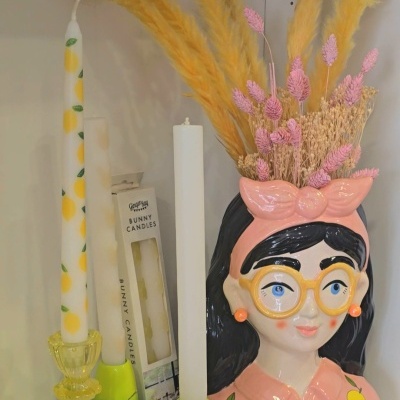 Vaso decorativo cerâmico com rosto feminino e flores secas, velas e embalagens ao lado.