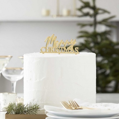 Bolo branco com topper dourado Merry Christmas em mesa decorada com árvore de Natal ao fundo