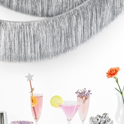 Quatro taças com bebidas rosa e decoração, franjas prateadas na parede, vaso branco com flor laranja