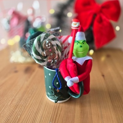 Caneca verde com pirulito, caneta com topo de Natal e boneco do Grinch em feltro numa mesa de madeira
