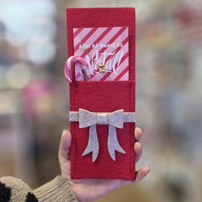 Embalagem vermelha de feltro com laço cinzento, cartão rosa, colar dourado e canequinho rosa e branco.