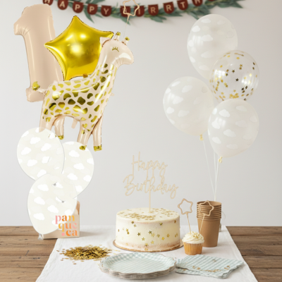 Decoração para festa de aniversário com balões dourados e brancos, bolo e pratos numa mesa de madeira