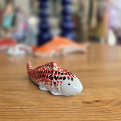 Peixe decorativo de cerâmica pintado de vermelho, preto e branco sobre mesa de madeira.
