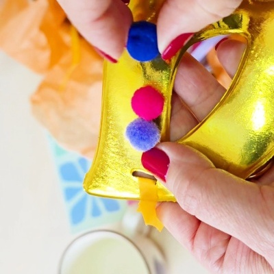 Mãos segurando objeto dourado em forma de letra com pompons coloridos