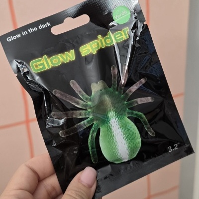 Embalagem preta brilhante com brinquedo verde de aranha, textos em inglês e português, mão segurando.