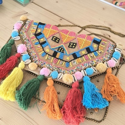 Bolsa colorida bordada com tasséis e conchas sobre superfície de madeira clara.